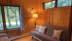 Ein gemütliches Blockhausszimmer mit Holzvertäfelung, einer Couch mit gemusterten Kissen, einer Lampe und Blick auf Bäume durch große Fenster.