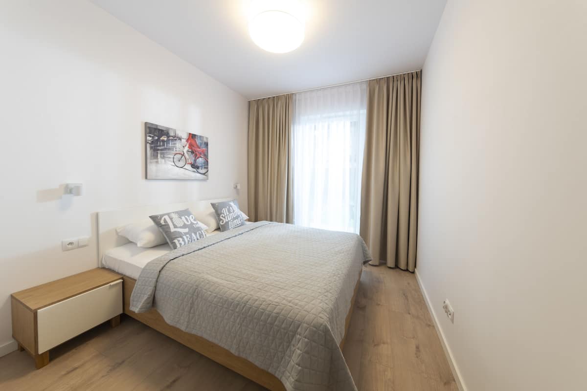 Un dormitorio con cama, mesita de noche, lámpara de pared, ventana con cortinas y un cuadro en la pared.