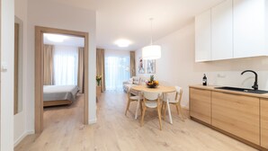 Une chambre d’hôtel moderne avec un plancher en bois, un coin repas avec une table ronde et des chaises, une cuisine avec un évier noir et un lit avec des rideaux beige.