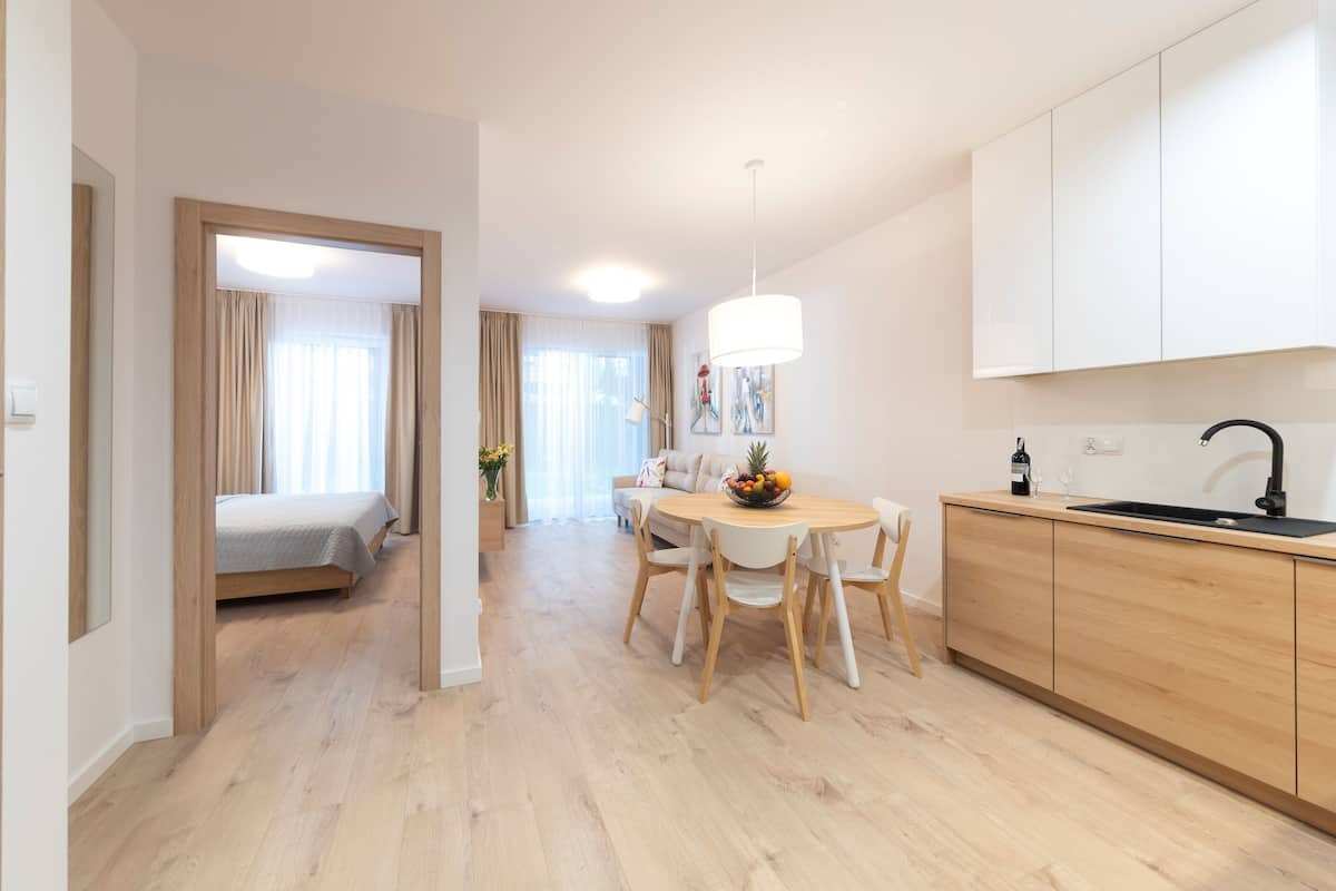 Una habitación de hotel moderna con piso de madera, un comedor con una mesa redonda y sillas, una cocina con un lavamanos negro, y una cama con cortinas color beige.