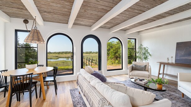 Amplia sala de estar con techo de madera, un sofá grande, una mesa de comedor con sillas y vistas a un campo a través de ventanas arqueadas.