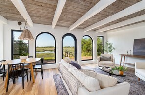 A spacious living room with a wooden ceiling, a large sofa, a dining table with chairs, and a view of a field through arched windows. - Marsh front villa on an acre. Private Dock + hot tub, steps from the beach  (St. Augustine)