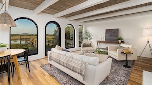 Amplia sala de estar con techo de madera, ventanas arqueadas grandes, un sofá blanco, un comedor con mesa y sillas de madera, y una pequeña mesita auxiliar con lámpara.