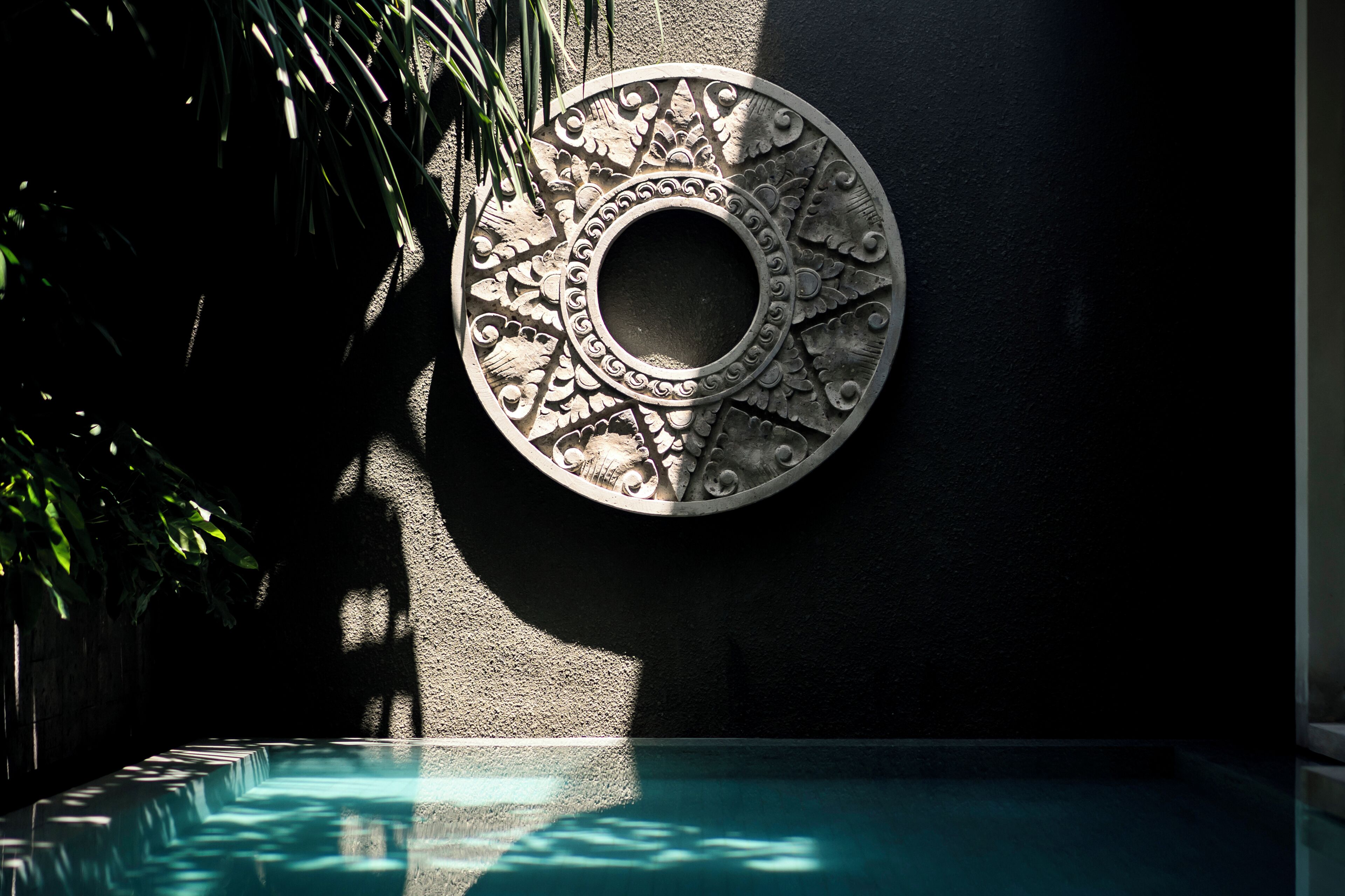 Un élément décoratif circulaire à accrocher au mur, orné de motifs complexes, une piscine et l’ombre d’une plante sur le côté gauche.