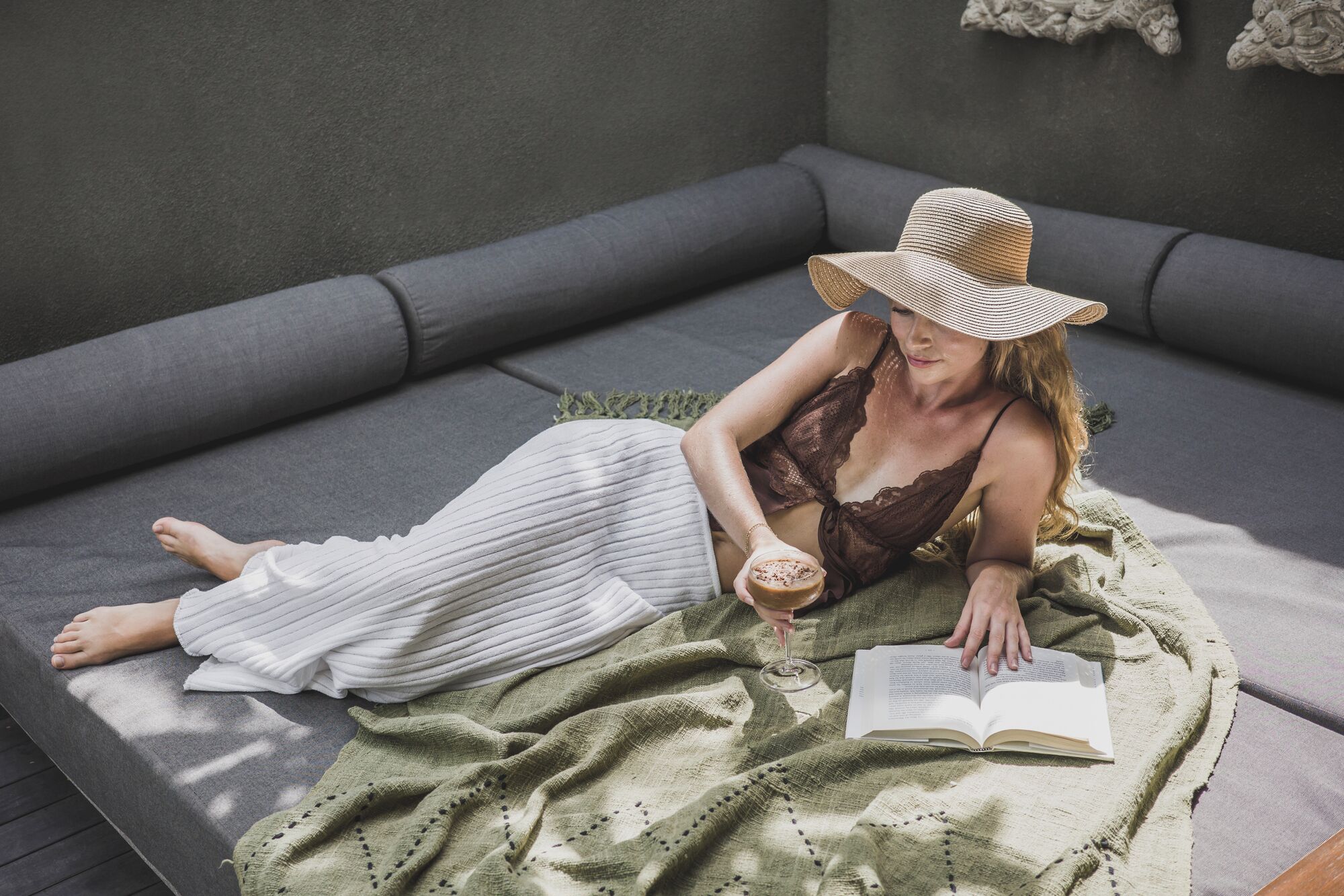 A person lying on a grey sofa with a green blanket, reading a book and holding a glass of drink.