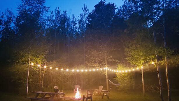An outdoor seating area with picnic tables and chairs, a campfire, and string lights in a wooded setting. - Secluded in the woods. No neighbors in sight. (Pullman)