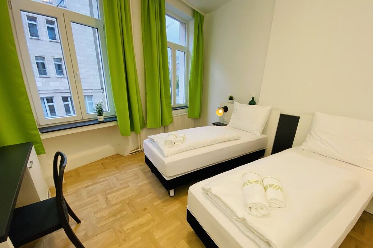 Une chambre d’hôtel avec deux lits, un bureau, une chaise, une fenêtre avec des rideaux et une table de chevet avec une lampe.