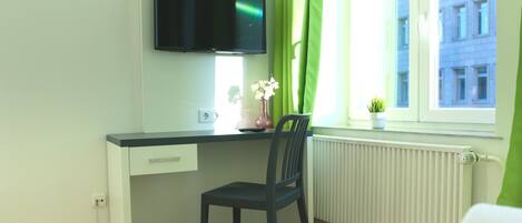 Una habitación con escritorio, silla, televisor, ventana con cortinas y radiador.
