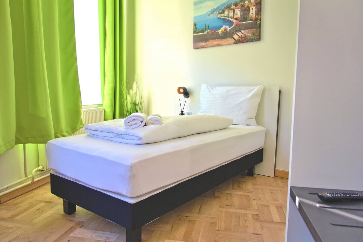 Un lit bien fait, avec un matelas et des oreillers blancs, une petite table de chevet sur laquelle se trouve une télécommande, et un tableau représentant un paysage côtier accroché au mur.