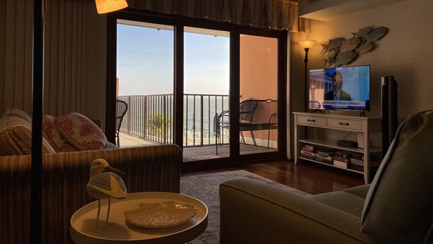 Una sala de estar con un sofá, una mesita con una escultura de un pájaro, un televisor sobre un mueble bajo, y un balcón con vista al mar.