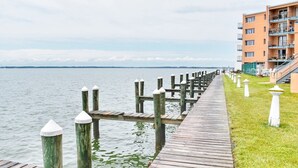 Un muelle de madera que se adentra en aguas tranquilas, con un área verde y un edificio con balcones a la derecha.