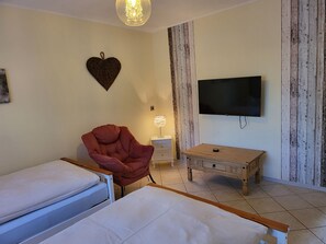 Un dormitorio con dos camas, una silla roja, una mesita de noche de madera con una lámpara, un televisor en la pared y un panel de pared de madera.