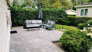 Un patio con mobiliario exterior gris, una sombrilla cubierta y un jardín bien cuidado con setos y flores.