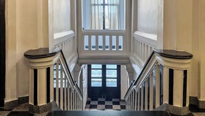 Un grand escalier, dont le sol est recouvert d’un motif à carreaux, mène à une vaste fenêtre en arc de cercle ornée de rideaux.