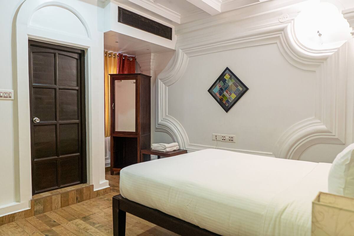 A bedroom with a bed, a door, a wall-mounted quilt, and a small table with a towel.