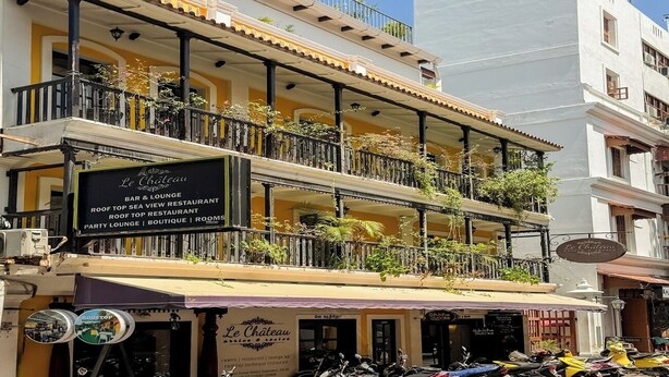 Un immeuble à plusieurs étages abritant un restaurant appelé « Le Château » au rez-de-chaussée, et doté de balcons ornés de plantes.