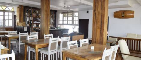A spacious dining area with wooden tables and white chairs, a bar area, and a bookshelf with various items.