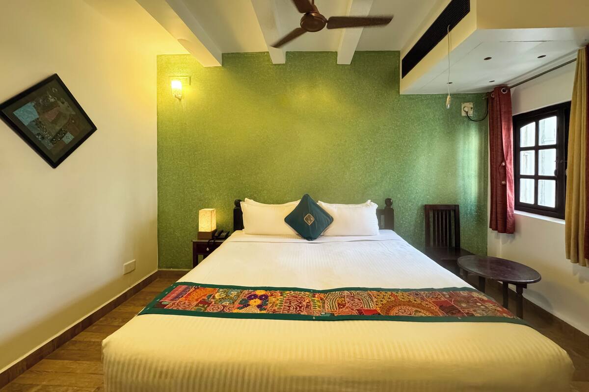 A hotel room with a large bed, a ceiling fan, a green accent wall, a window with curtains, and a nightstand with a lamp.