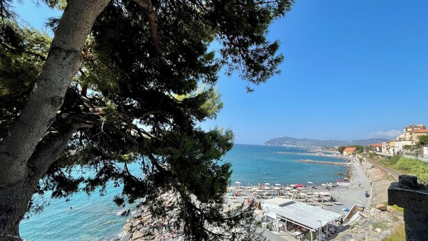 A coastal view with a rocky beach, a clear blue sea, and a few buildings in the background. - Piccola Navetta (Diano San Pietro)