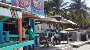 Un puesto de comida en la playa con un letrero colorido, bancos de madera y una zona a la sombra.