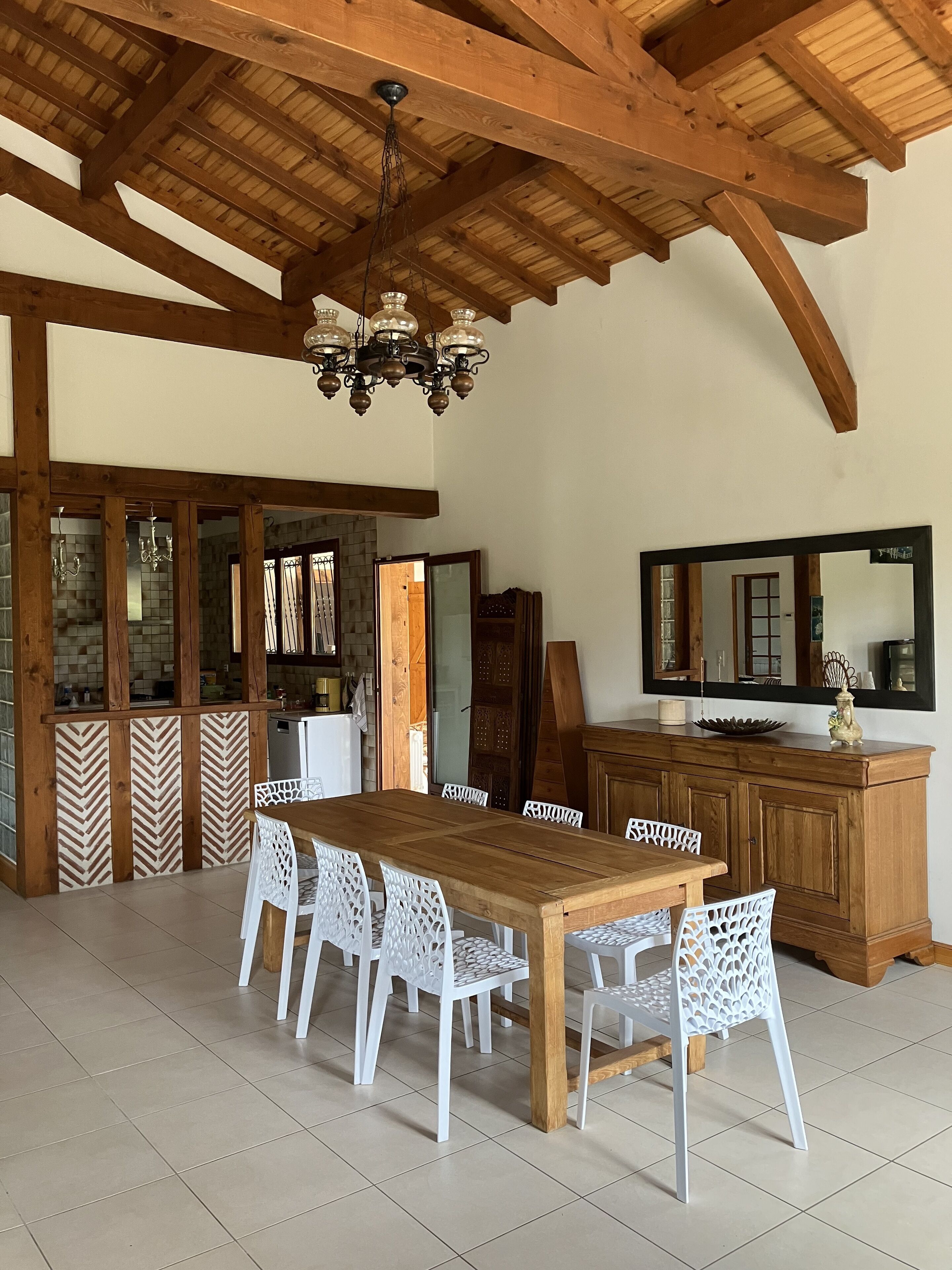 A dining area with a wooden table and white chairs, a chandelier, and a wooden cabinet with a mirror.