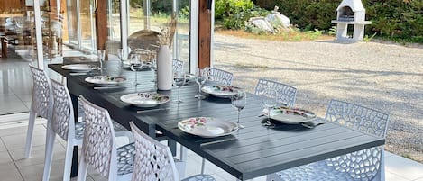 An outdoor dining area with a covered patio, a long table set for six, and a stone oven in the background.