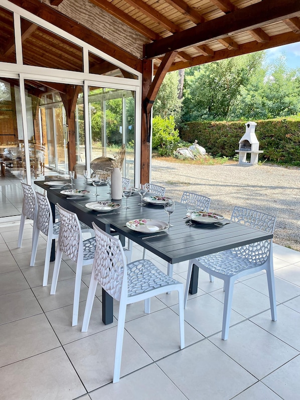 Een overdekt terras met een eethoek, een lange tafel gedekt voor zes personen en een stenen oven op de achtergrond.