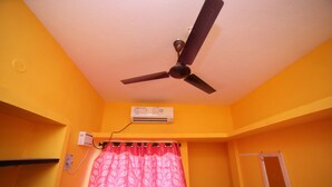 Una habitación con ventilador de techo, aire acondicionado, una cortina floral rosa y un ventanal pequeño.