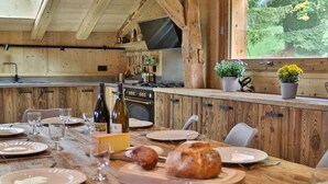 Un intérieur rustique en bois, avec une longue table à manger dressée pour un repas, un coin cuisine équipé d’une cuisinière et de différents ustensiles suspendus, et une fenêtre donnant sur un paysage verdoyant.