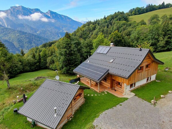 Een houten chalet met een schuin dak en zonnepanelen, omringd door weelderige begroeiing en bergen.