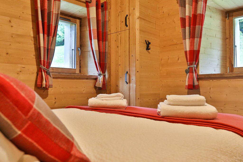 Una habitación tipo cabaña de madera con una cama, dos ventanas con cortinas y una puerta pequeña.