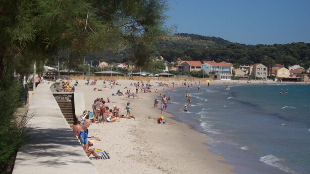 A beach with people sunbathing and swimming, a concrete walkway, and buildings in the background.