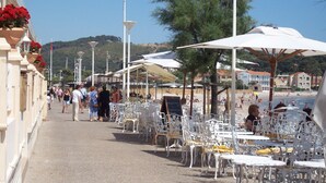 A seaside café with outdoor seating, white metal chairs, and umbrellas.