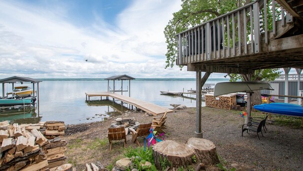 Une terrasse au bord du lac, avec des bancs en bois et un espace abrité, des bateaux amarrés et une pile de bois de chauffage.