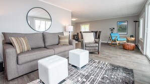 Un salon avec un canapé gris, un pouf blanc et un tapis à motifs. On y trouve un fauteuil à bascule et une petite table avec une lampe.