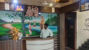 A reception desk with a man standing behind it, a mural with a rural scene, and a ceiling with circular light fixtures. - Hotel Prakash (Kilvelur)