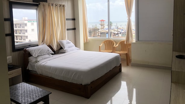 A hotel room with a large bed, a small table, a chair, and a balcony with a view of the sea and buildings. - Hotel Prakash (Kilvelur)