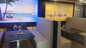 A restaurant with a large TV screen displaying a beach scene, cushioned seating, and a wall with a family-themed poster. - Hotel Prakash (Kilvelur)