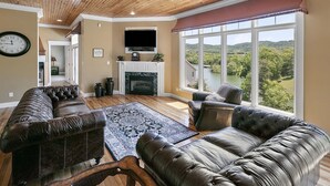 A spacious living room with a fireplace, leather sofas, a large window with a view of a lake, and a clock on the wall.