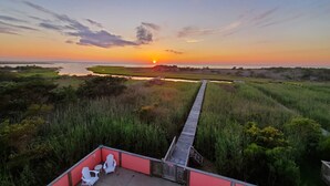 A sunset view from a deck with a pool, chairs, and a wooden walkway leading to a beach. - Amazing Sound Front, Ocean View Home with Pool, Spa and Dock (Frisco)