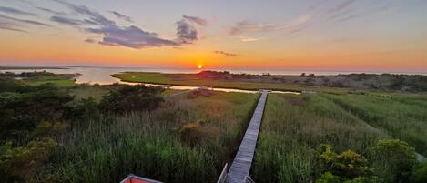 A sunset view from a deck with a pool, chairs, and a wooden walkway leading to a beach.