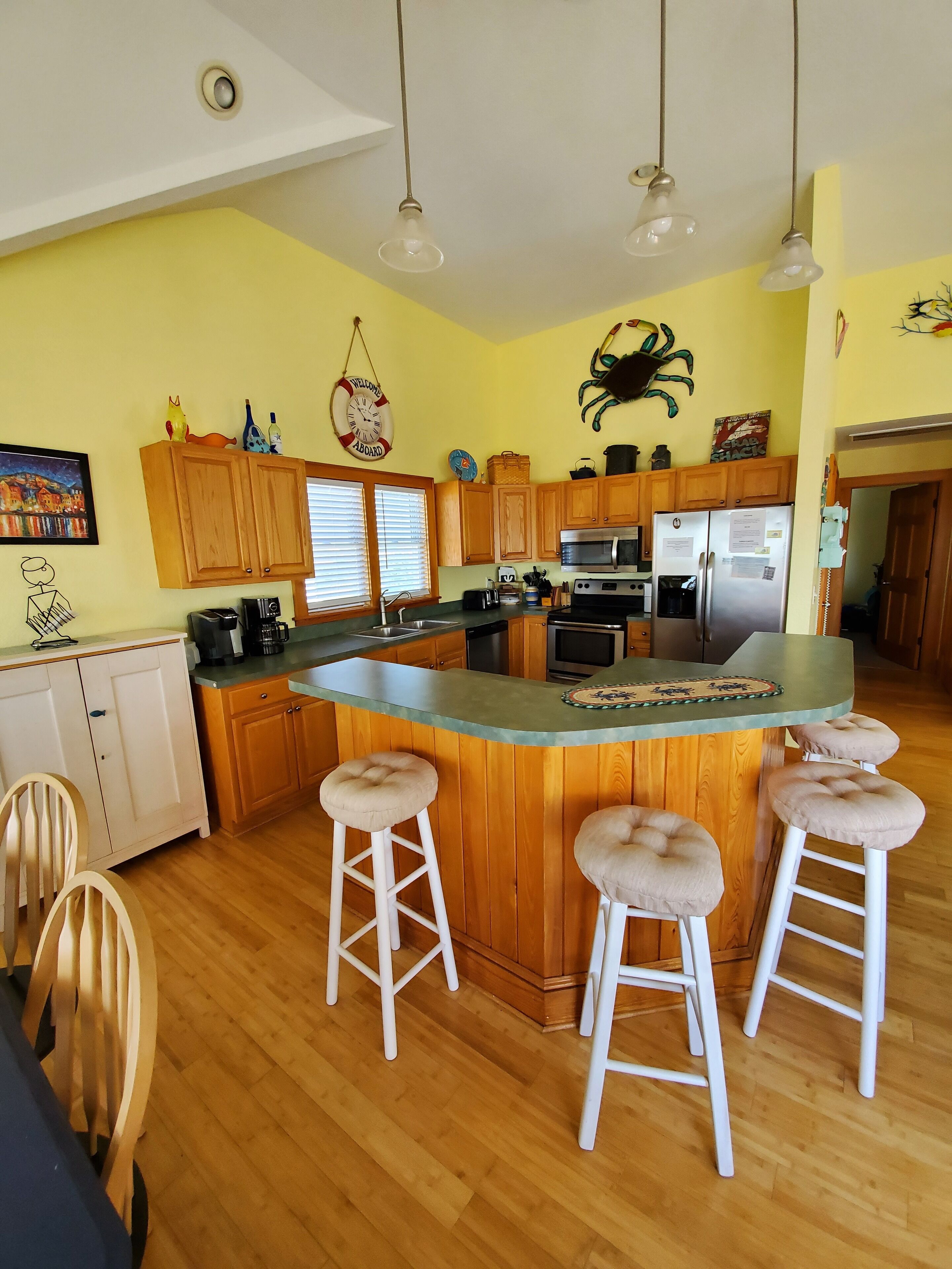 A kitchen with wooden cabinets, a central island with stools, stainless steel appliances, and a "Welcome" sign on the wall.