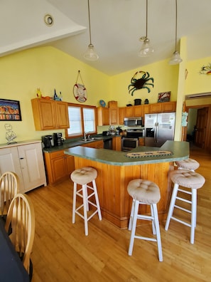 A kitchen with wooden cabinets, a central island with stools, stainless steel appliances, and a "Welcome" sign on the wall. - Amazing Sound Front, Ocean View Home with Pool, Spa and Dock (Frisco)