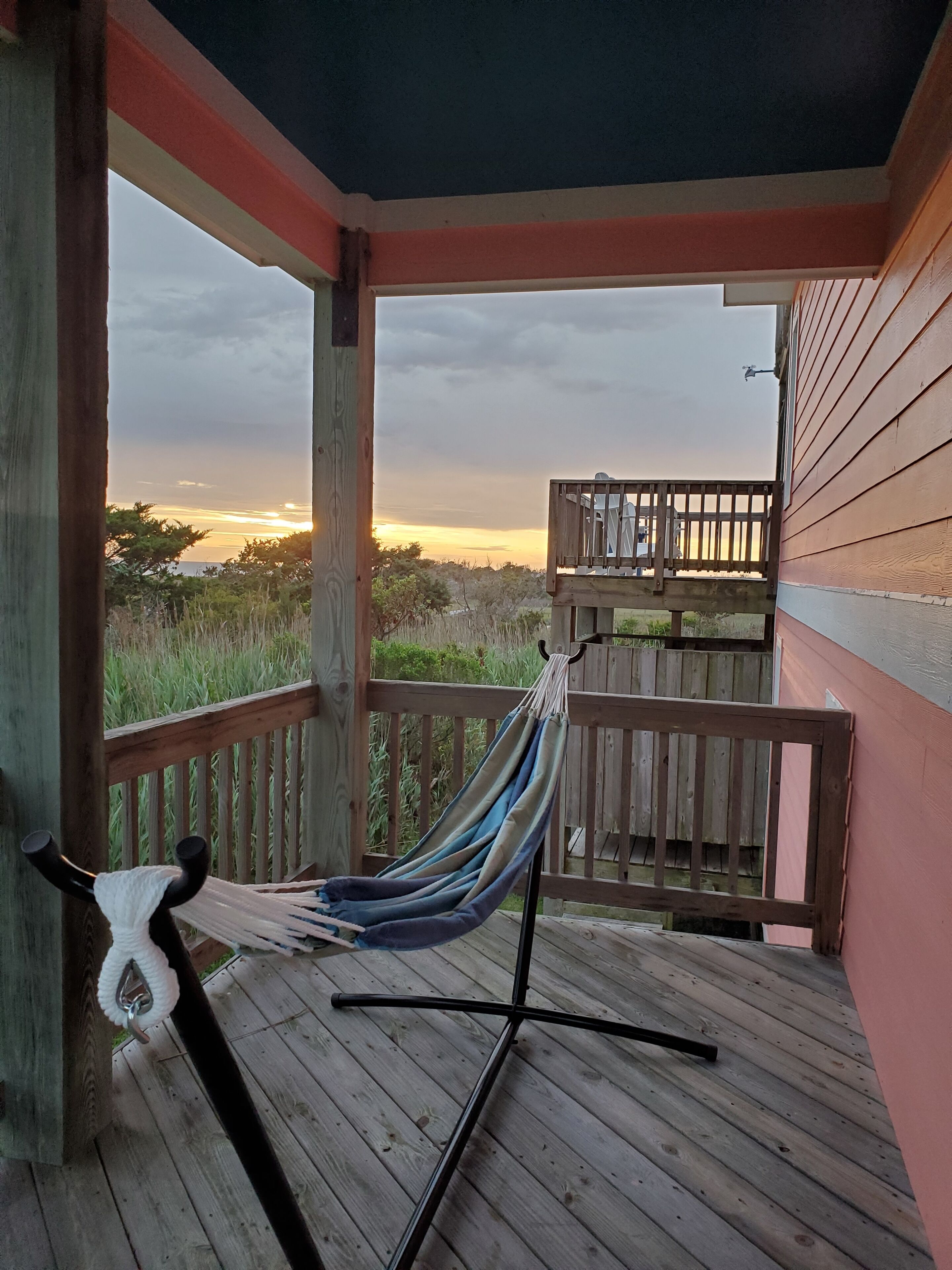 A wooden deck with a hammock and a view of the sunset over a grassy landscape.