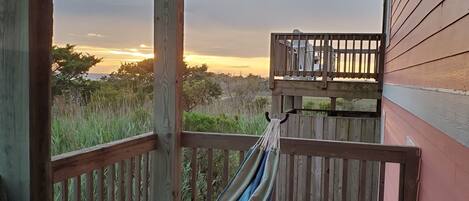 A wooden deck with a hammock and a view of the sunset over a grassy landscape.