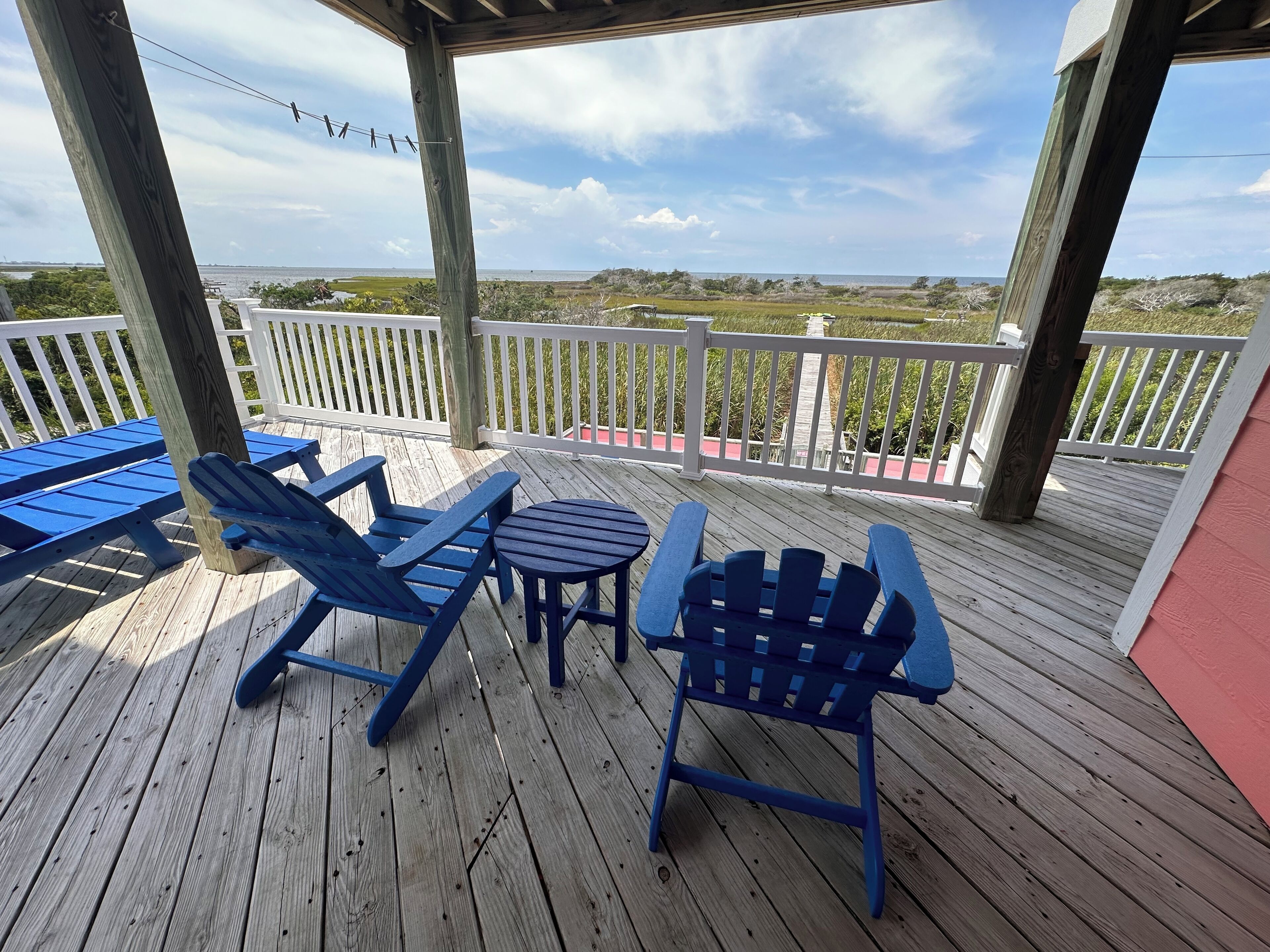 A wooden deck with blue Adirondack chairs and a table, overlooking a coastal landscape.