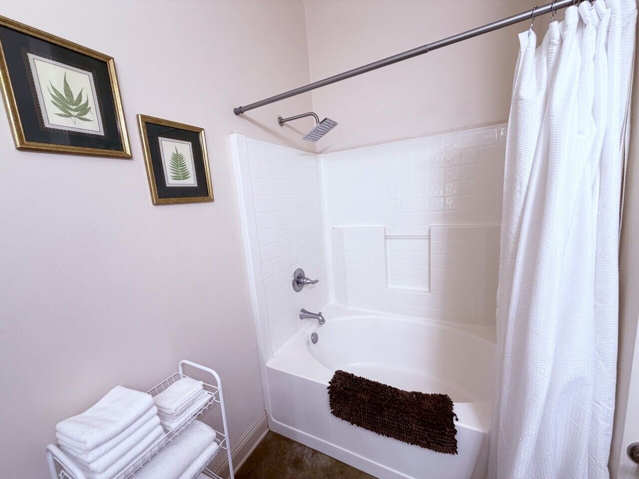 A bathroom with a bathtub, a shower, a towel rack with towels, and two framed botanical prints on the wall.