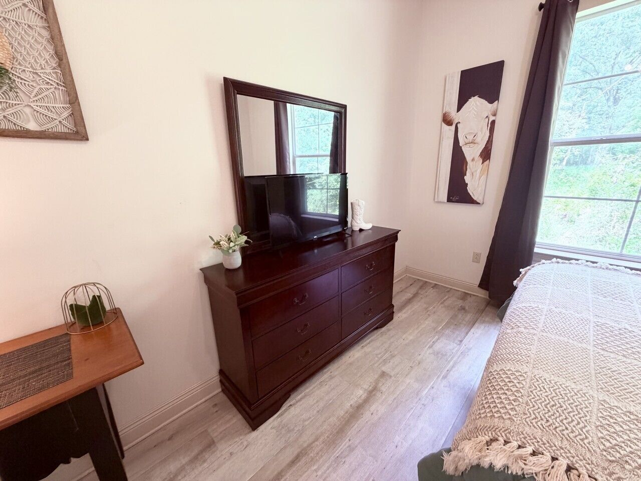 A bedroom with a wooden dresser, a television, a bed with a textured blanket, and a window with curtains.