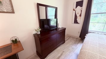 Een slaapkamer met een houten dressoir, een televisie, een bed met een gestructureerde deken en een raam met gordijnen.