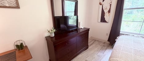 A bedroom with a wooden dresser, a television, a bed with a textured blanket, and a window with curtains.